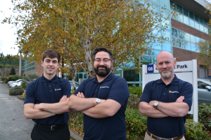 Three male employees of Modern Plant, wearing navy blue collared shirts with the company logo, stand outside the office building with their arms crossed. The building has glass and brick elements, and there are trees with autumn foliage behind them.